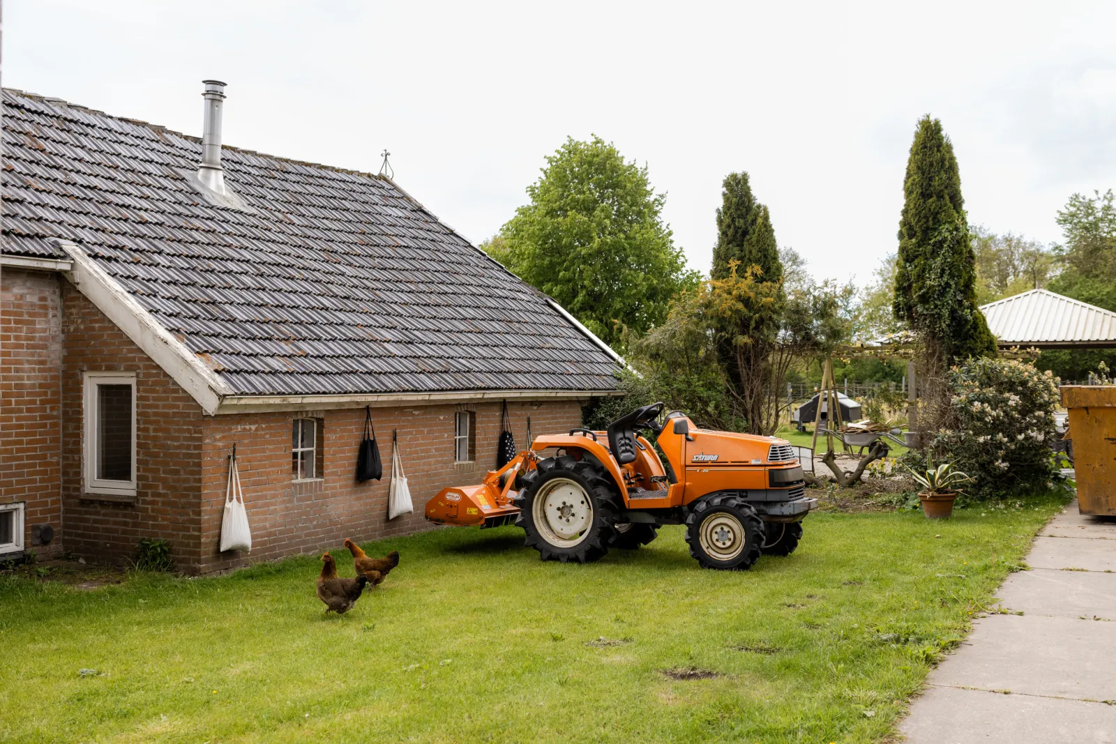 Een huis met een landbouwwerktuig erbij geparkeerd. Twee kippen lopen over het gras.